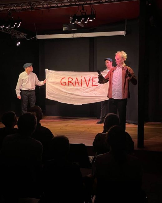 Trois comédiens sur scène tenant une banderole « GRAIVE »